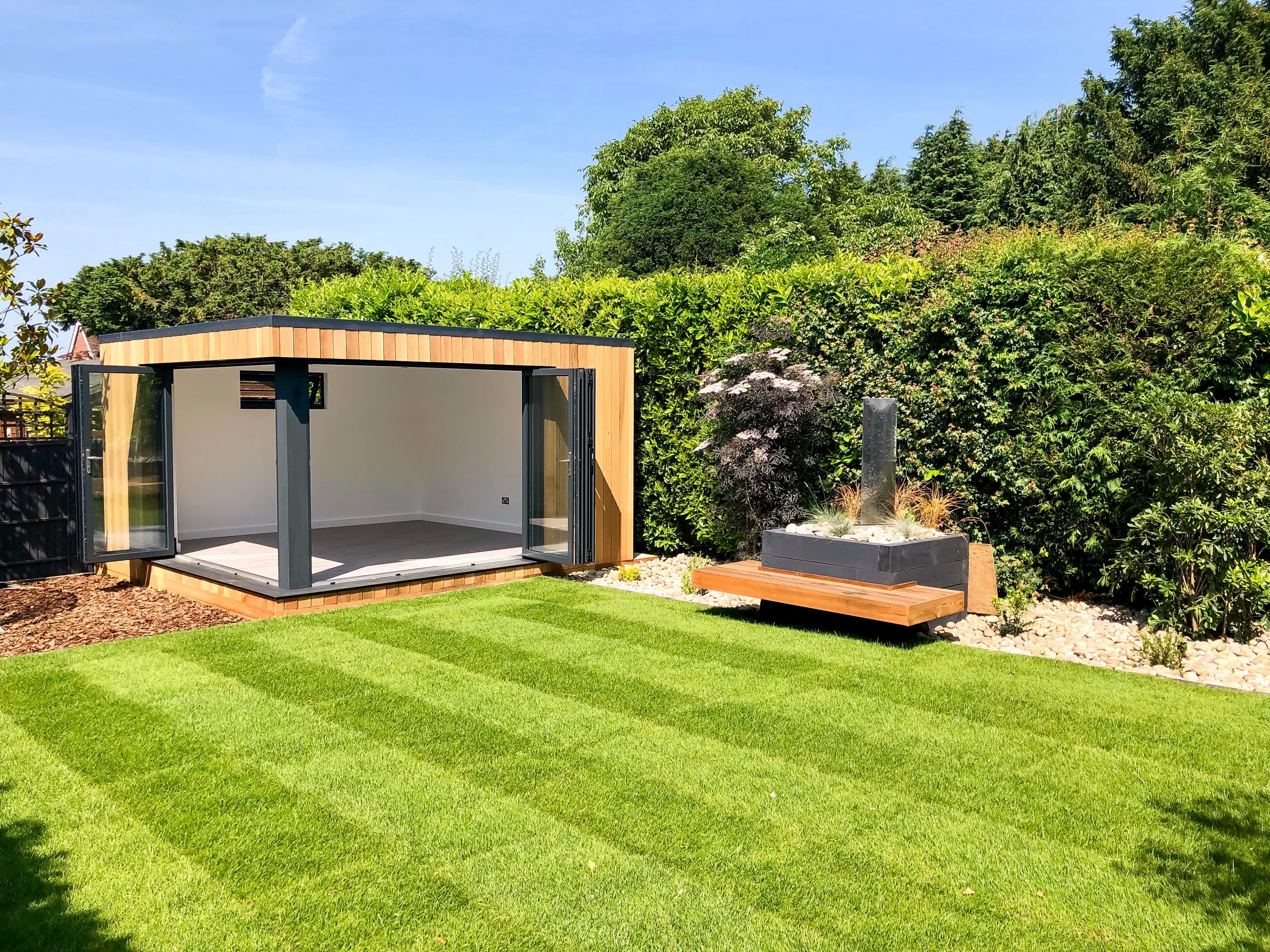 Exterior day view of a small Vivid Green garden office with open anthreacite grey aluminium bi-fold doors, surrounded by tall hedges and grass, with a wooden bench to the right