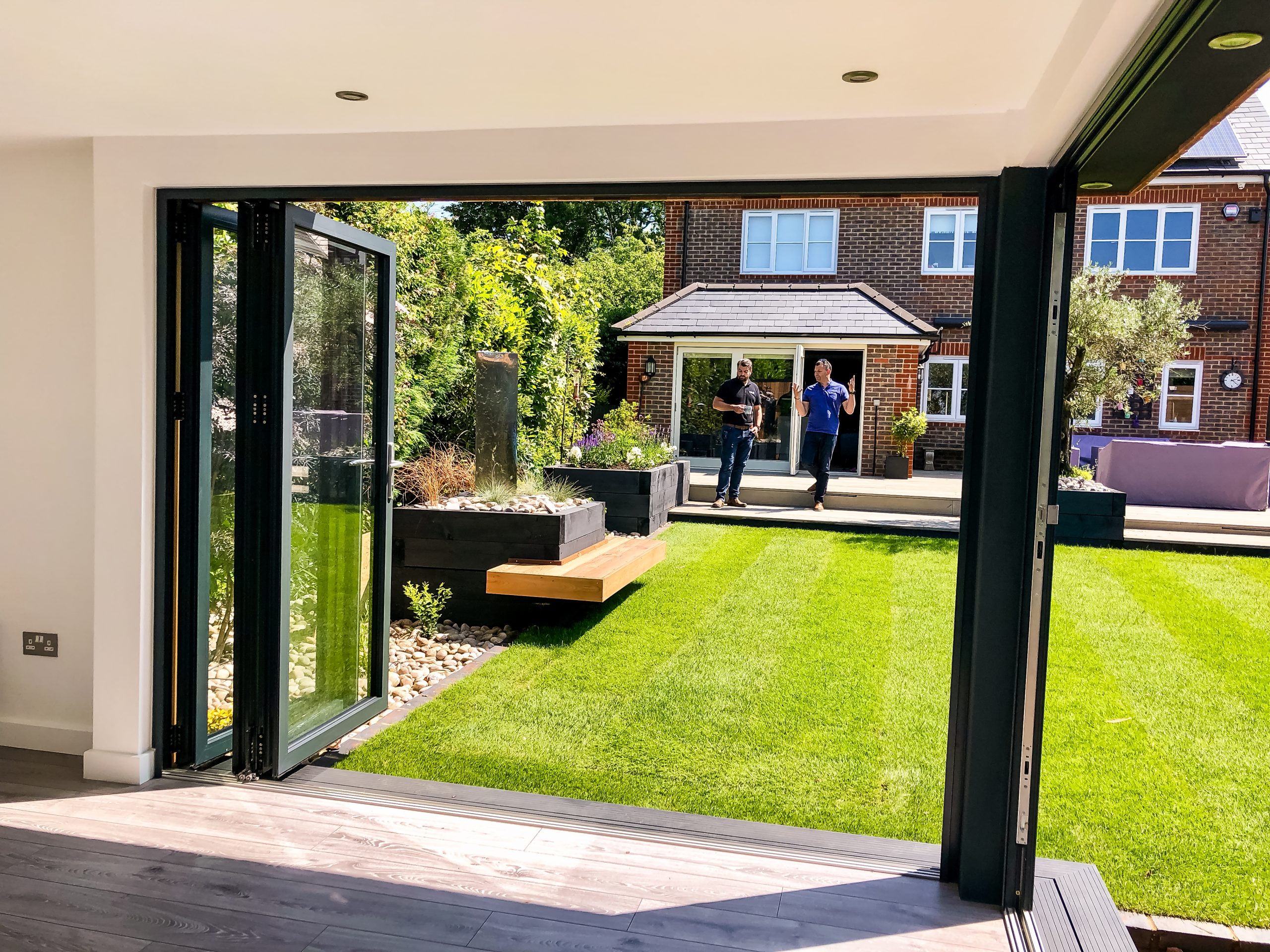Interior view of small unfurnished Vivid Green garden room with anthracite grey framed corner bi-fold doors which open onto the garden with a wooden bench to the left of the garden and the main house with two people standing in the background