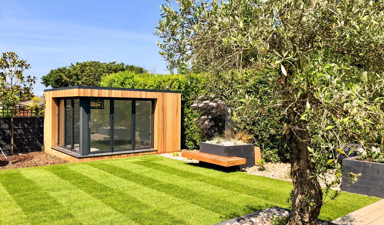 Exterior day view of a small Vivid Green garden office with closed anthracite grey framed corner glass bi-fold doors, surrounded by tall hedges and grass, with a wooden bench to the right and a tree in the foreground