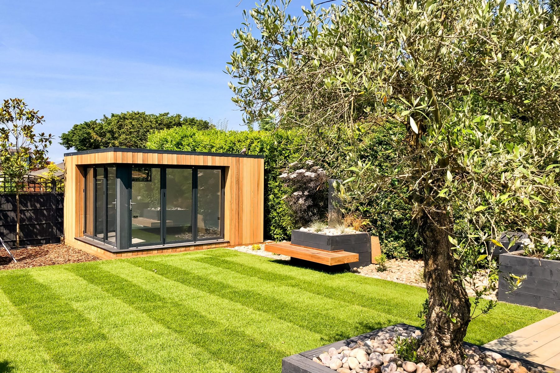 Exterior day view of a small Vivid Green garden office with closed anthracite grey framed corner glass bi-fold doors, surrounded by tall hedges and grass, with a wooden bench to the right and a tree in the foreground