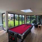 Interior view of a garden games room featuring anthracite grey bi-fold doors and windows with a skylight in the roof, furnished with a red and black pool table and an office desk in the corner, providing a view of the garden