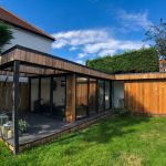 Vivid Green L-shaped garden office space with western red cedar cladding and anthracite grey aluminium doors and windows, slightly elevated above the garden with an outside patio covered by an open overhang with decked seating area.