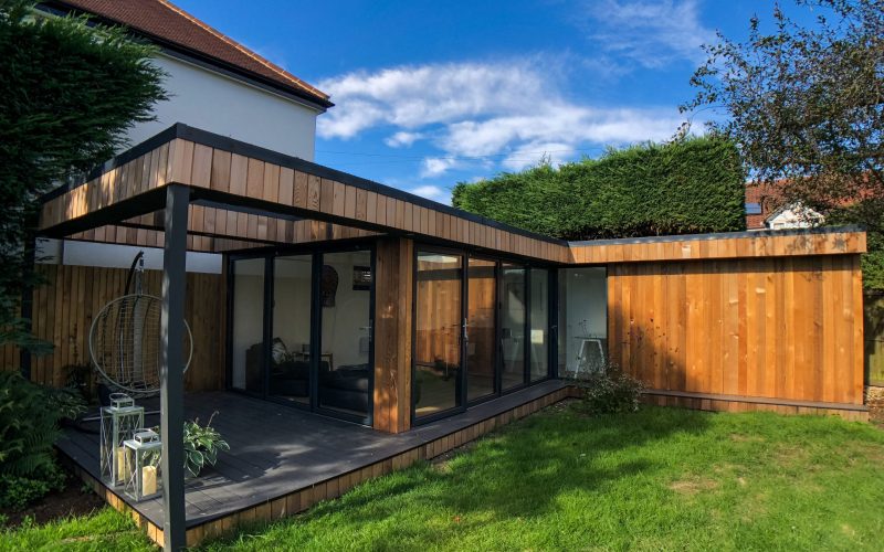 Vivid Green L-shaped garden office space with western red cedar cladding and anthracite grey aluminium doors and windows, slightly elevated above the garden with an outside patio covered by an open overhang with decked seating area.