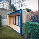 Vivid Green garden office with larch rain screen cladding, grey aluminium finishes, and anthracite grey bi-fold doors situated in a small lush garden with an overhang at the entrance and a brick wall to the left.