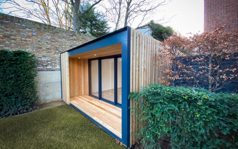 Vivid Green garden office with larch rain screen cladding, grey aluminium finishes, and anthracite grey bi-fold doors situated in a small lush garden with an overhang at the entrance and a brick wall to the left.