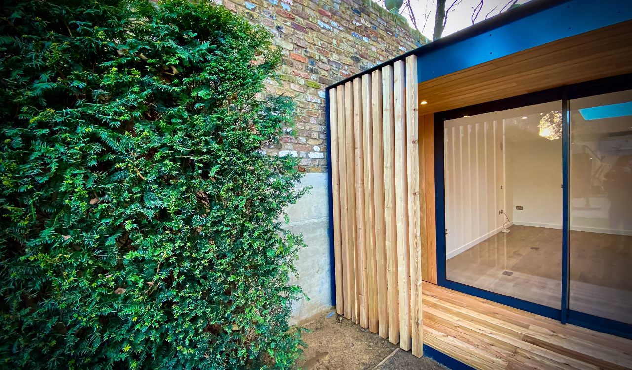 Exterior view of Vivid Green garden office space with larch rain screen cladding and anthracite grey aluminium sliding doors, featuring a slight overhang above the entrance framed in aluminium fascia, situated next to a green hedge and brick neighboring wall