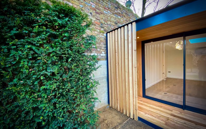 Exterior view of Vivid Green garden office space with larch rain screen cladding and anthracite grey aluminium sliding doors, featuring a slight overhang above the entrance framed in aluminium fascia, situated next to a green hedge and brick neighboring wall