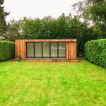 Exterior view of a Vivid Green garden office space with anthracite grey aluminium bi-fold doors and windows, large decking area and flat roof, situated at the bottom of the garden flanked by two tall hedges.