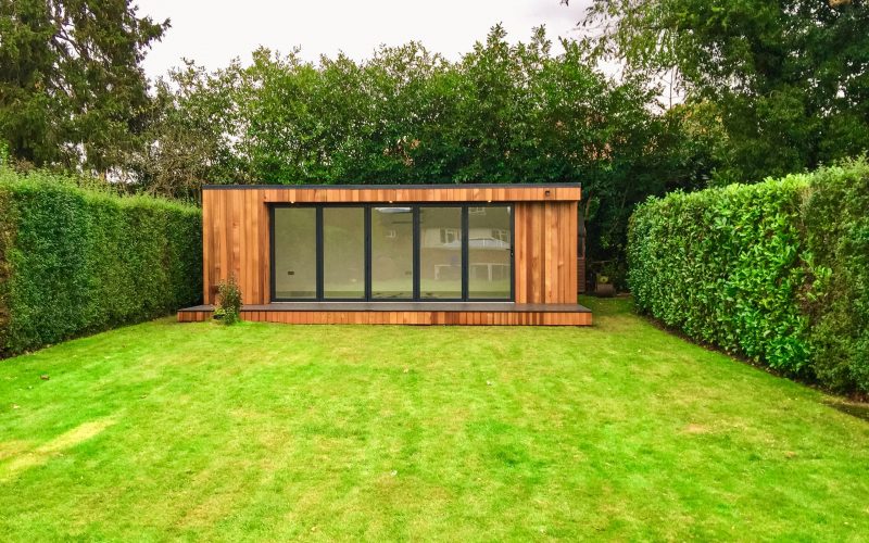Exterior view of a Vivid Green garden office space with anthracite grey aluminium bi-fold doors and windows, large decking area and flat roof, situated at the bottom of the garden flanked by two tall hedges.