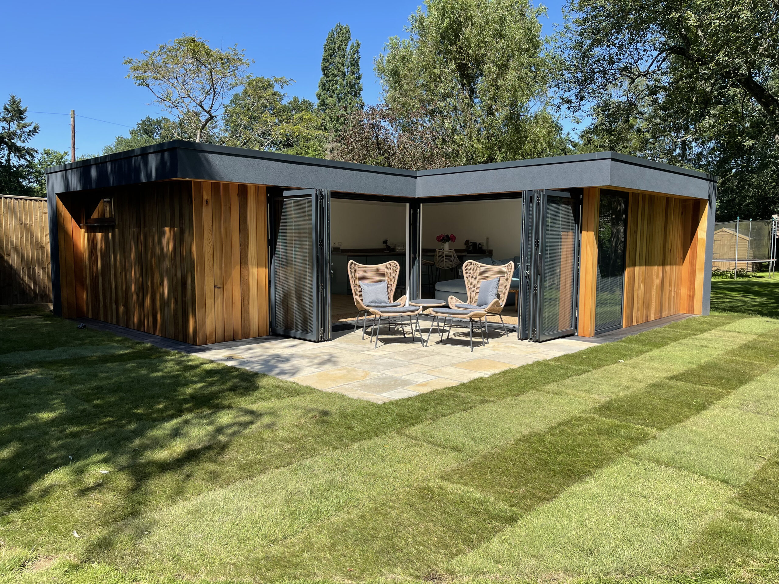 Exterior view of a Vivid Green L-shaped western red cedar clad garden studio with an anthracite grey rendered overhang and flat roof, anthracite grey aluminium bi-fold doors opening onto a small tiled patio, and furniture set on it, surrounded by an artificial grass garden on a sunny day.