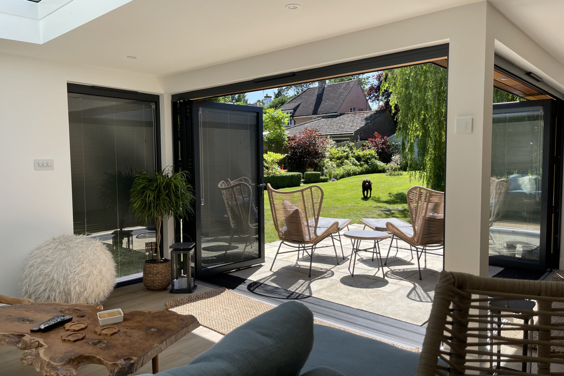 Interior of a Vivid Green furnished L-shaped garden room with anthracite grey aluminium bi-fold doors with integral blinds opening onto a tiled patio with outdoor furniture and a sunny grass lawn, with the main house visible in the background.