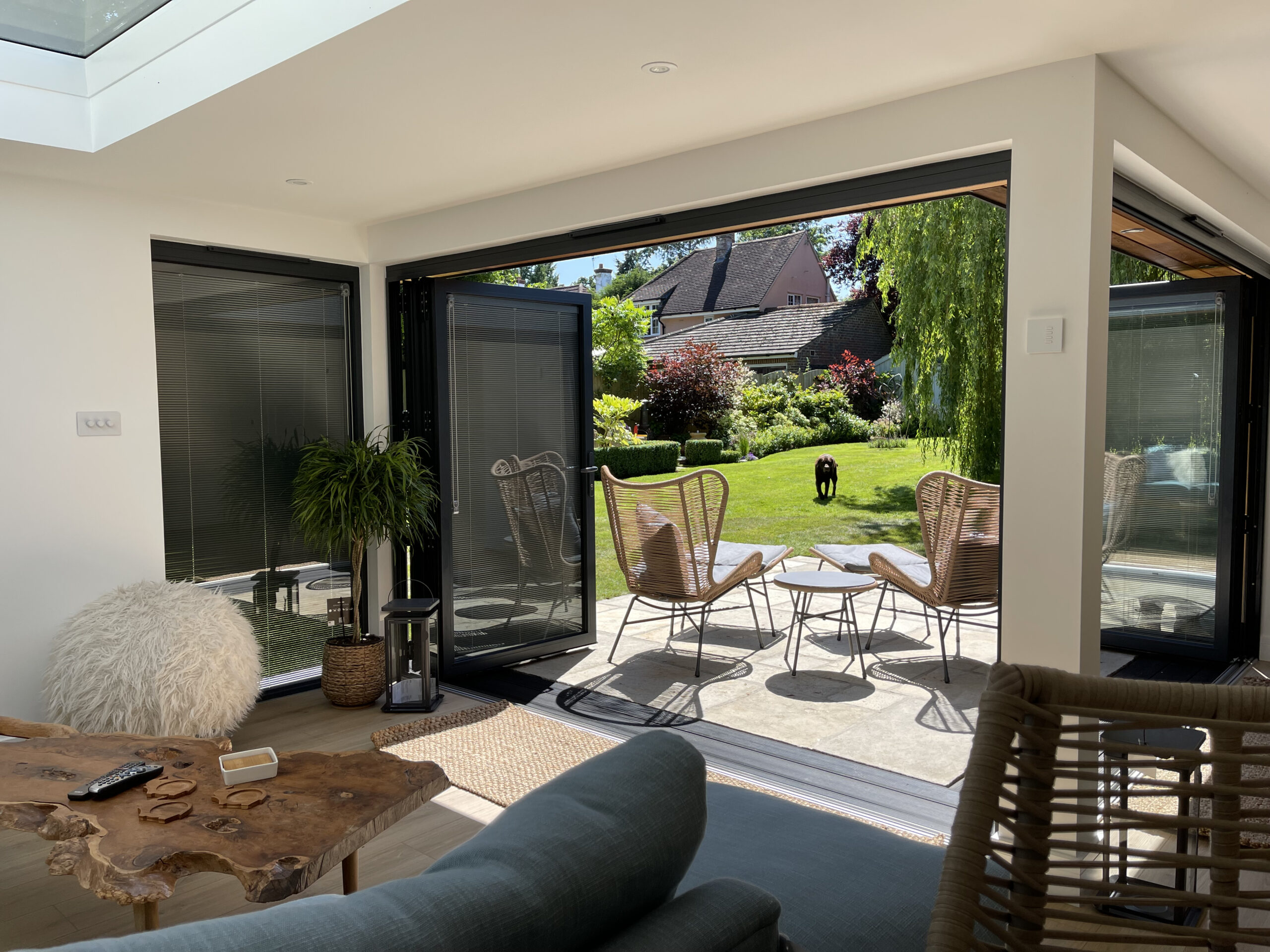 Interior of a Vivid Green furnished L-shaped garden room with anthracite grey aluminium bi-fold doors with integral blinds opening onto a tiled patio with outdoor furniture and a sunny grass lawn, with the main house visible in the background.