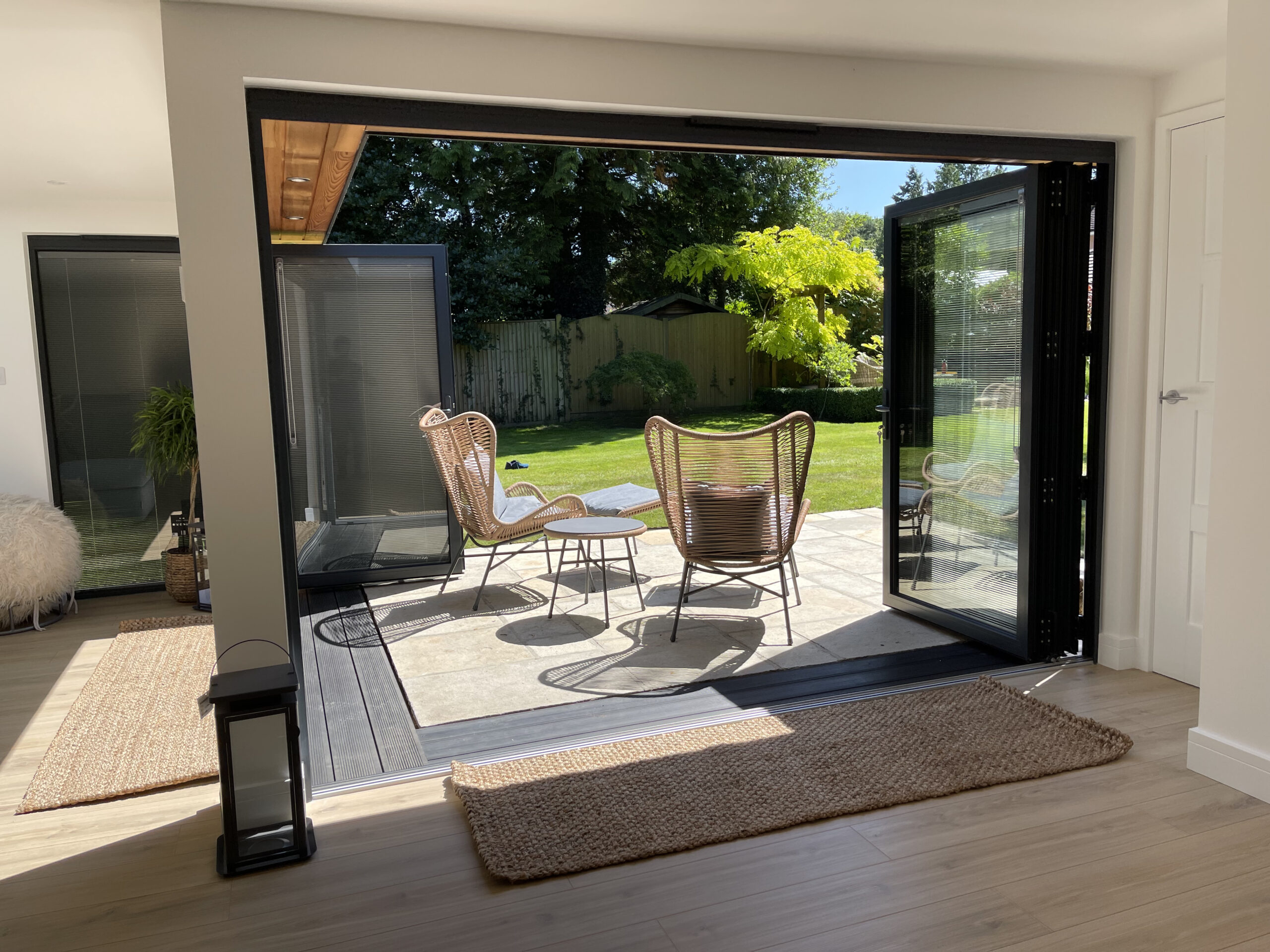 Interior view of a Vivid Green furnished L-shaped garden room with open anthracite grey aluminium bi-fold doors, wooden floors with rugs, and outdoor tiled patio with garden furniture overlooking a sunny grass lawn.