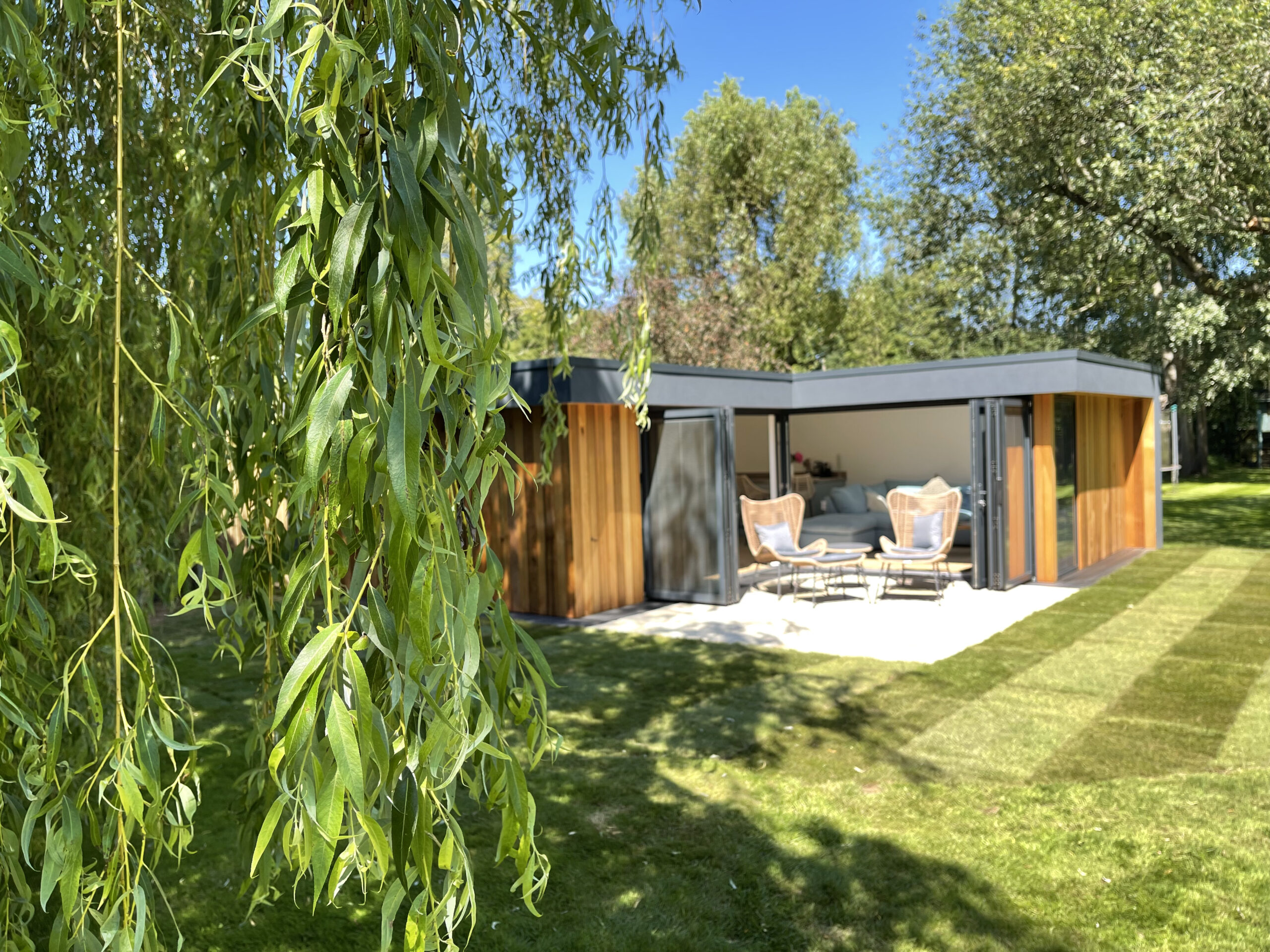 Exterior view of a Vivid Green flat roof L-shaped garden studio with western red cedar cladding on the walls and anthracite grey render framing the overhang, open anthracite grey aluminium bi-fold doors and windows, and a small outdoor furniture set on a tiled patio surrounded by grass and trees on a sunny day
