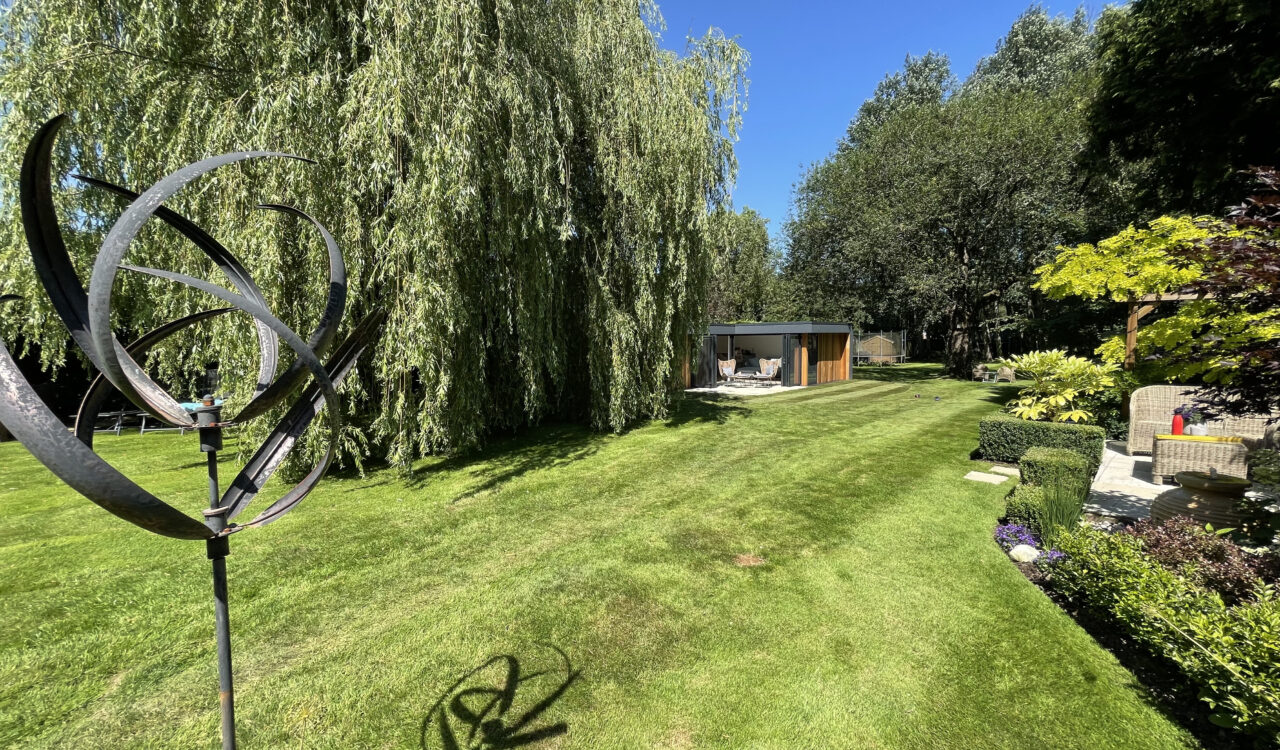 Long shot of a sunny garden with a metal ornament in the foreground, a willow tree and outdoor seating area in the middle ground, and a Vivid Green L-shaped western red cedar clad garden room in the background.