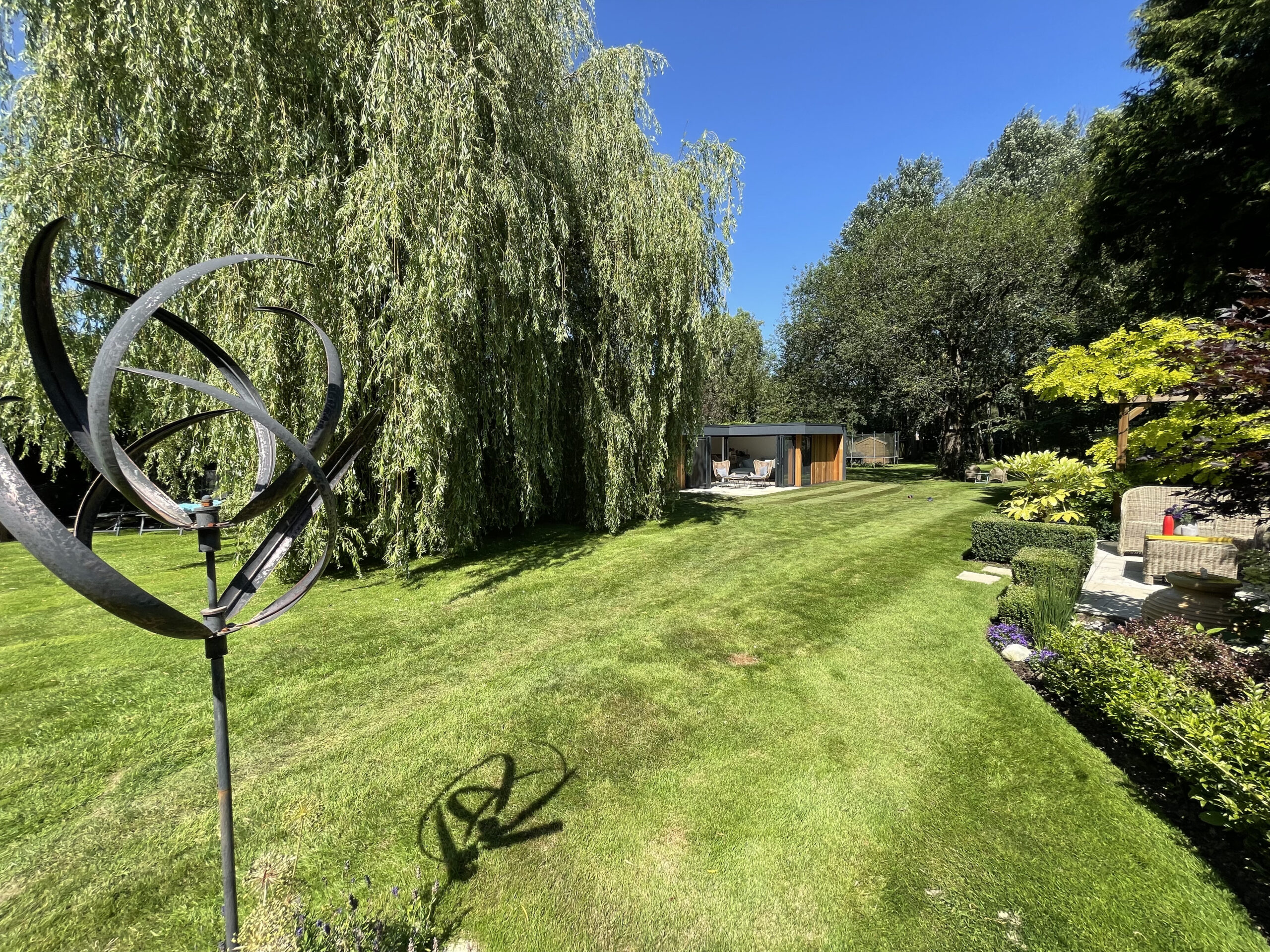 Long shot of a sunny garden with a metal ornament in the foreground, a willow tree and outdoor seating area in the middle ground, and a Vivid Green L-shaped western red cedar clad garden room in the background.