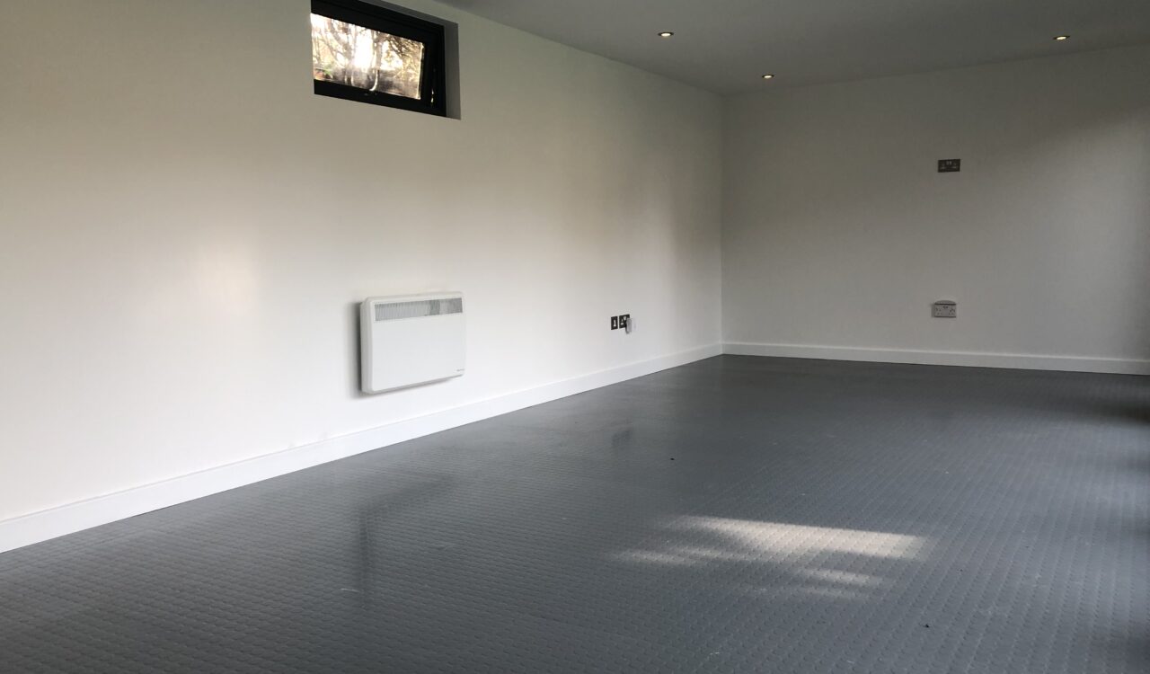 Unfurnished Vivid Green family garden room interior with grey rubber tiled flooring, recessed lights, white walls with electrical outlets, panel heater under a small black-framed window and a TV wall mount.