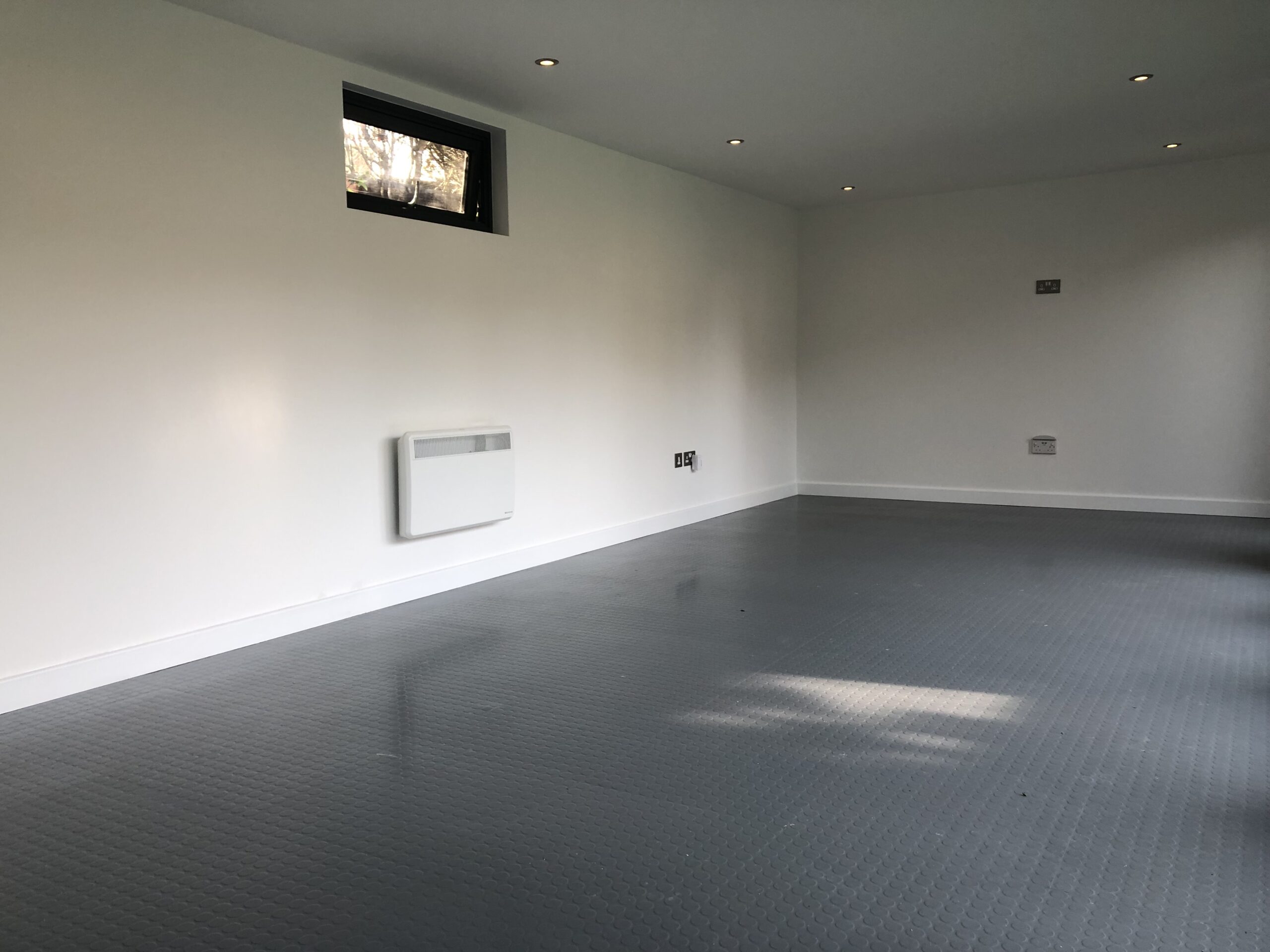Unfurnished Vivid Green family garden room interior with grey rubber tiled flooring, recessed lights, white walls with electrical outlets, panel heater under a small black-framed window and a TV wall mount.