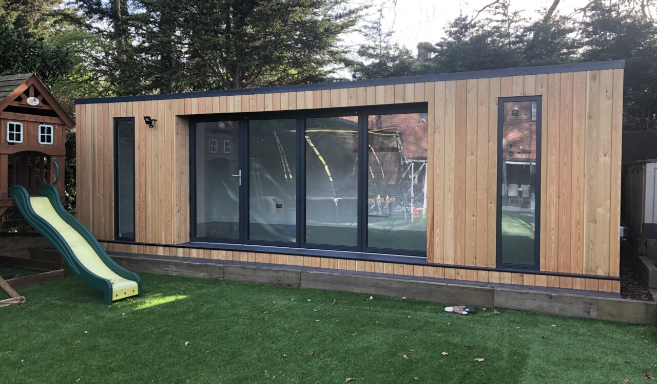 Exterior view of a larch clad Vivid Green flat-roofed family garden room with anthracite grey windows and aluminium bi-fold doors opening to artificial grass, featuring a playhouse and slide to the left and a trampoline reflected in the windows.