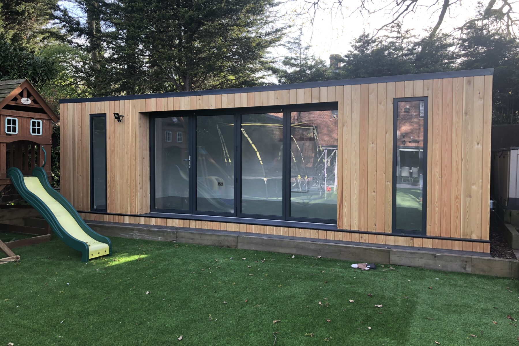 Exterior view of a larch clad Vivid Green flat-roofed family garden room with anthracite grey windows and aluminium bi-fold doors opening to artificial grass, featuring a playhouse and slide to the left and a trampoline reflected in the windows.