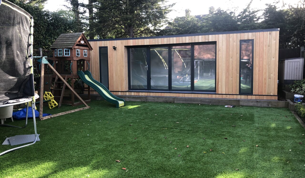 Vivid Green, larch clad, flat-roofed family garden room with anthracite grey windows and aluminium bi-fold doors opening to a backyard featuring artificial grass, trampoline, and a playhouse with a slide for kids.