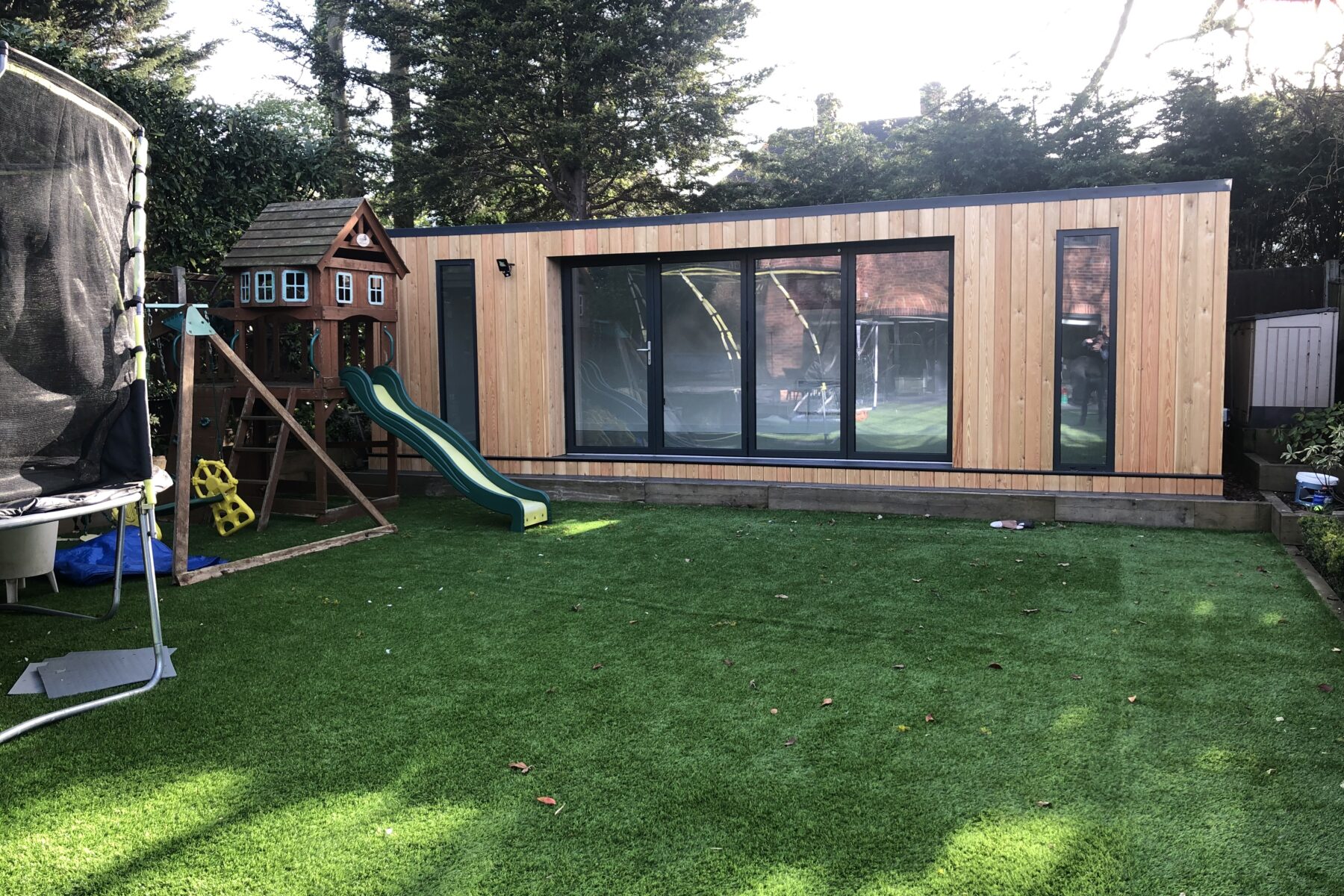 Vivid Green, larch clad, flat-roofed family garden room with anthracite grey windows and aluminium bi-fold doors opening to a backyard featuring artificial grass, trampoline, and a playhouse with a slide for kids.
