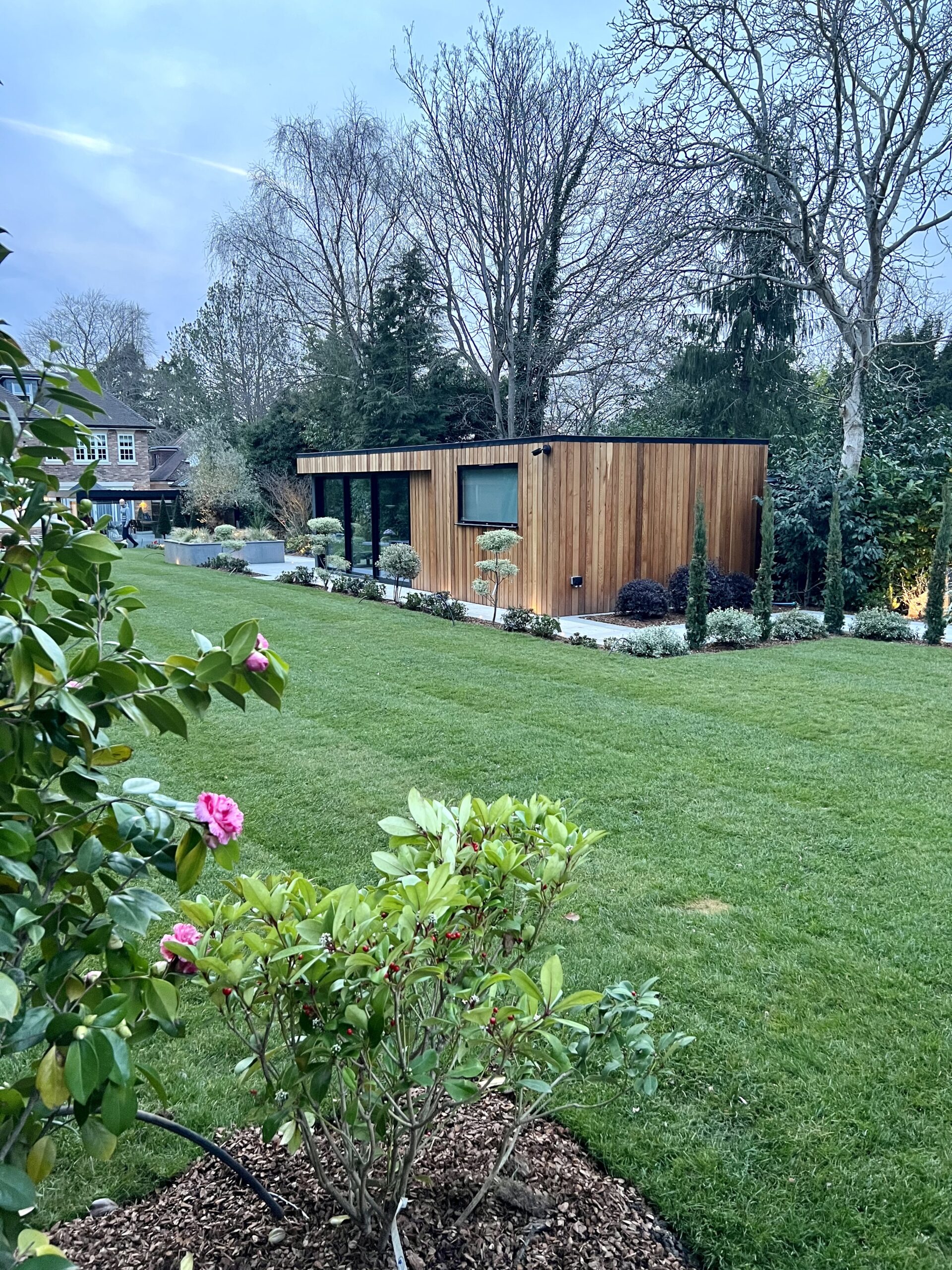 Exterior view of a lush garden featuring a flat roofed, western red cedar clad, Vivid Green garden studio with grey framed windows and doors and an overhang with grey finishes and trees in the background