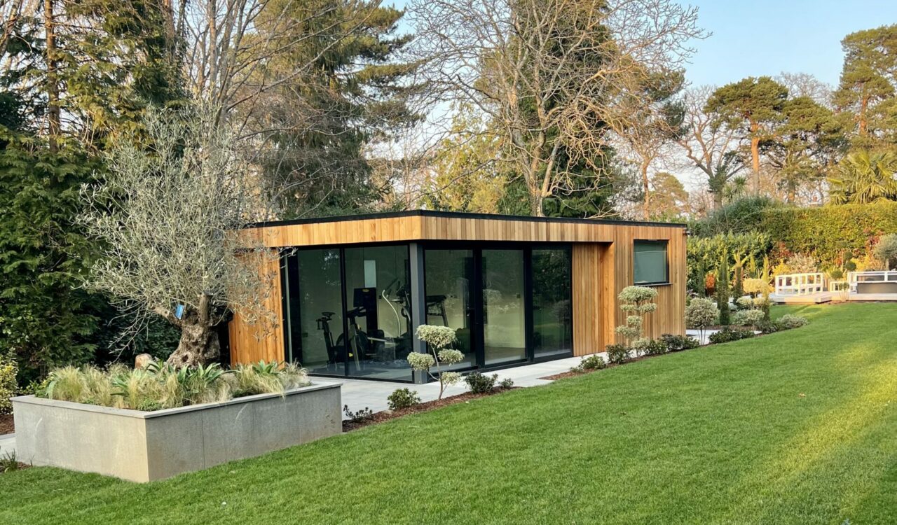 Exterior view of a Vivid Green garden gym with western red cedar cladding, a flat roof, aluminium grey framed corner glass sliding doors, surrounded by a tiled patio, trees in the background, and lush foliage