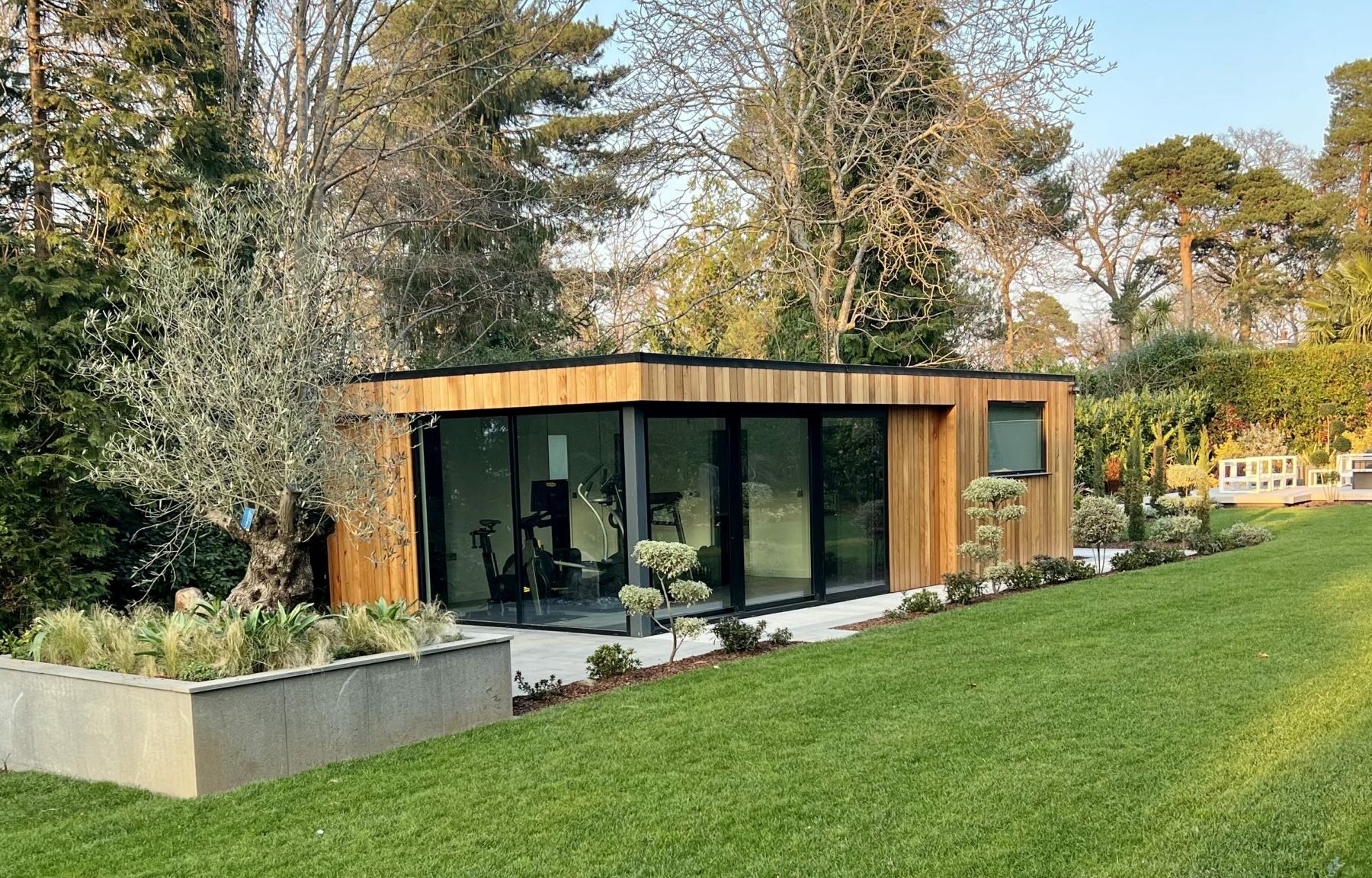 Exterior view of a Vivid Green garden gym with western red cedar cladding, a flat roof, aluminium grey framed corner glass sliding doors, surrounded by a tiled patio, trees in the background, and lush foliage