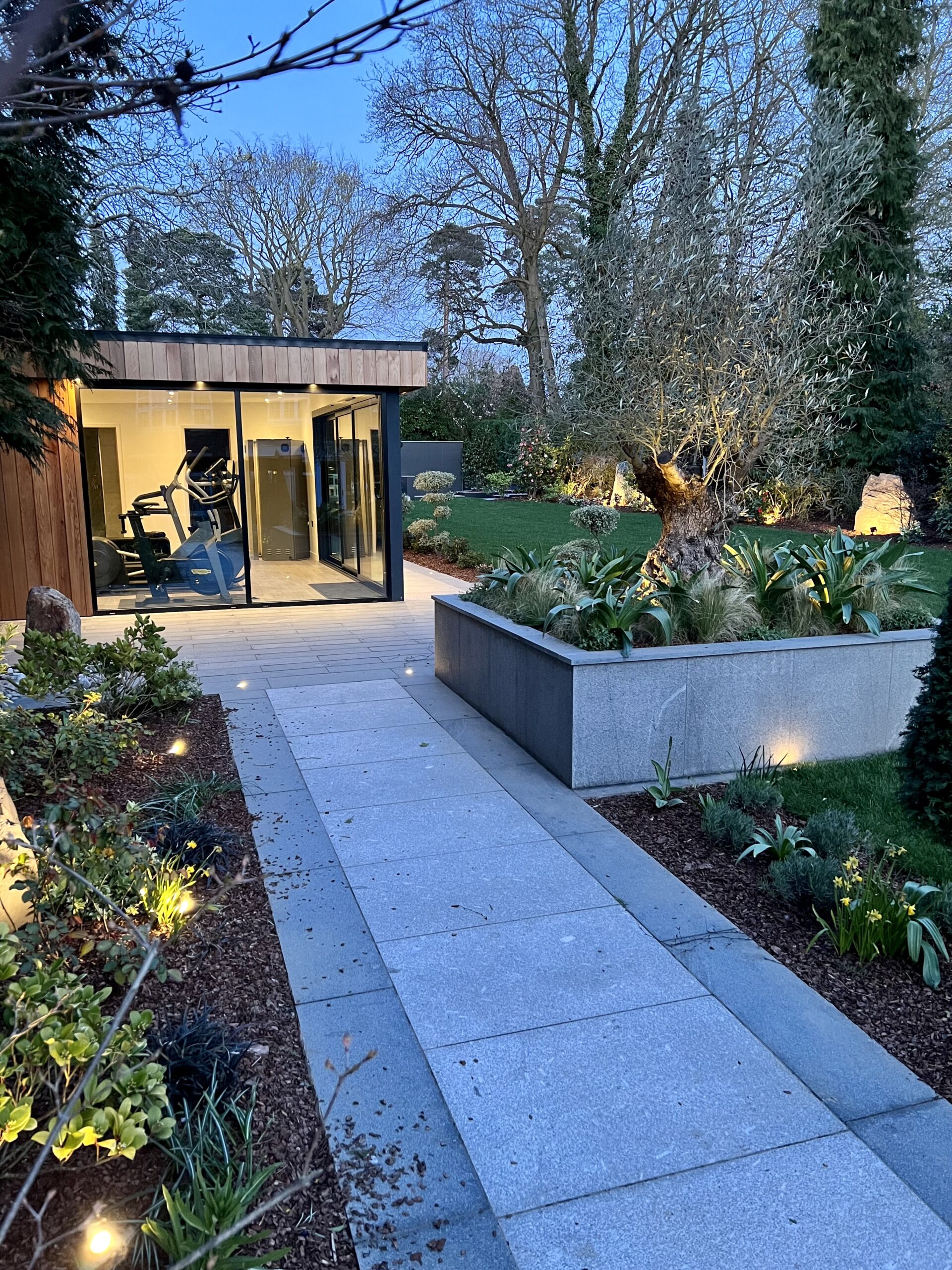 Evening exterior view of a Vivid Green garden gym featuring anthracite grey aluminium corner glass sliding doors, surrounded by garden foliage, with a concrete walkway leading to the entrance and trees in the background.