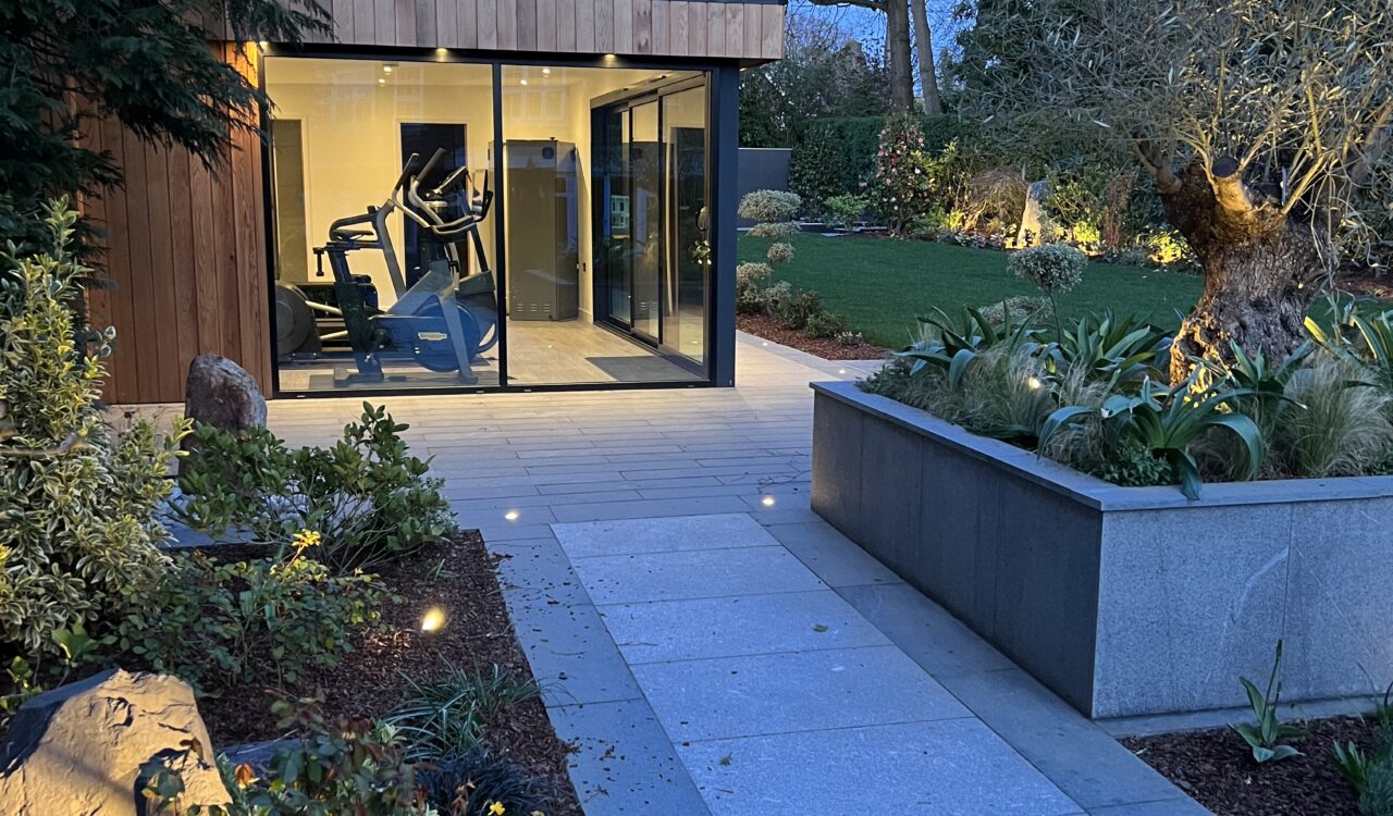 Evening exterior view of a Vivid Green garden gym studio featuring cedar cladding, anthracite grey framed corner glass sliding doors, surrounded by garden foliage, with a concrete walkway leading to the entrance and trees in the background.
