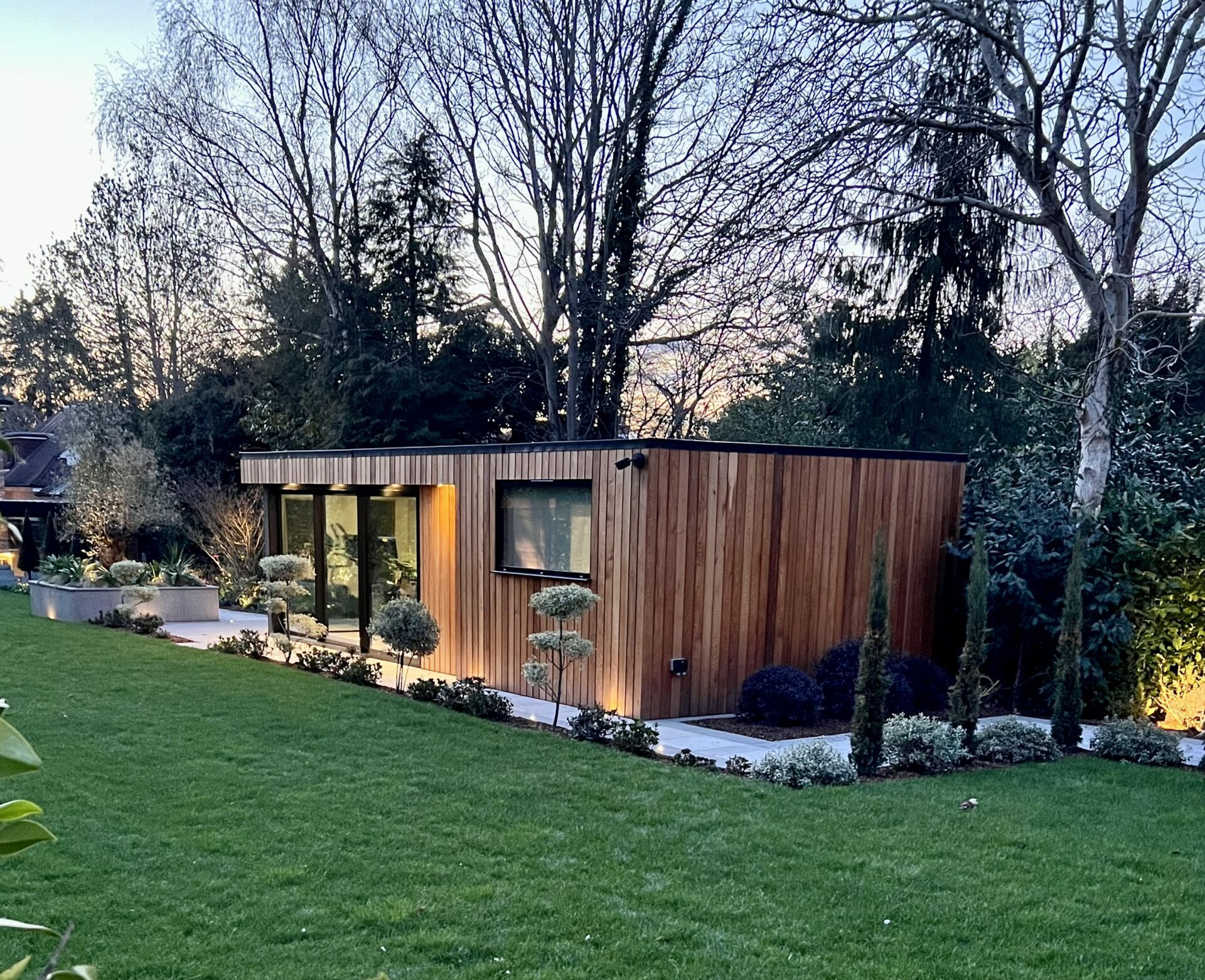Exterior evening view of a lush garden featuring a flat roofed, western red cedar clad, Vivid Green garden studio with a anthracite grey framed window and an overhang with grey finishes and trees in the background