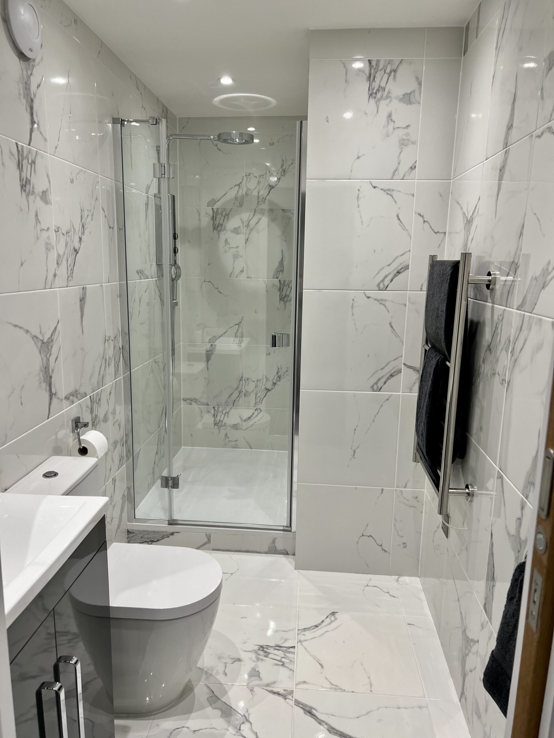 Interior shot of a brightly lit Vivid Green garden studio shower room with toilet featuring marble tiling on the floor and walls, silver bathroom fittings a shower with a frameless glass door, a white toilet and the corner of the bathroom sink