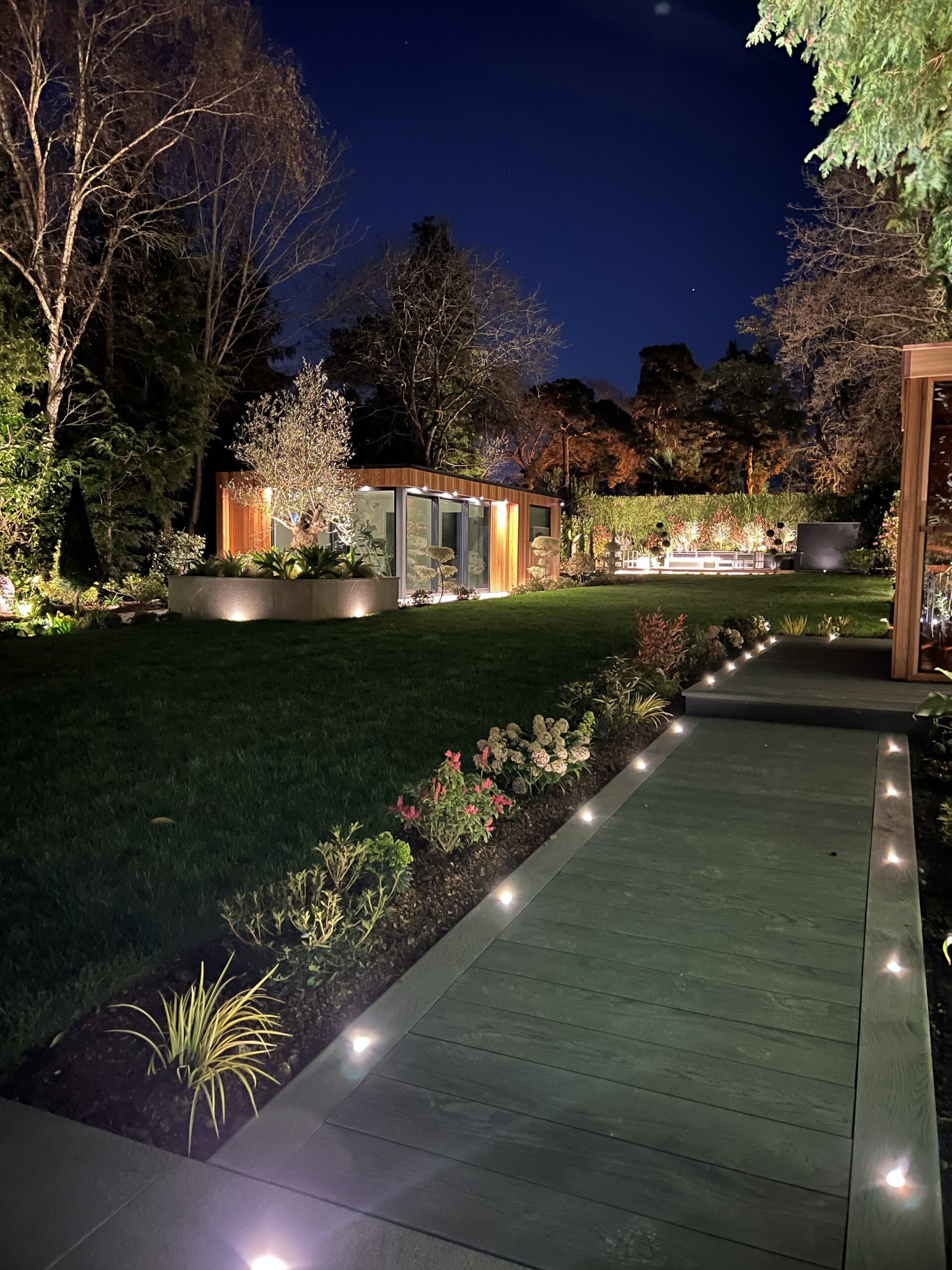 Exterior night view of a wood walkway in a garden leading up to a Vivid Green garden studio with cedar cladding a grey door and in the background across the garden to the right a smaller garden studio with a connected patio and furniture