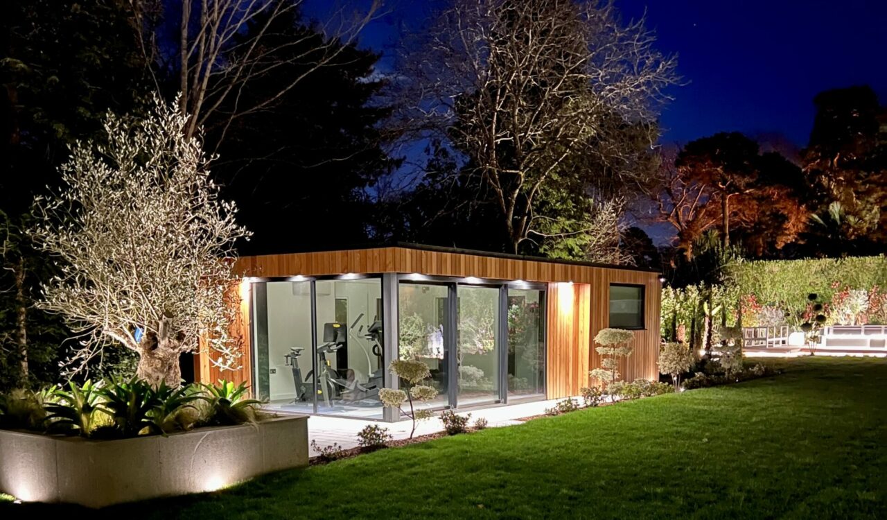 Exterior evening view of aVivid Green garden gym lit up by canopy lights, grey framed corner glass sliding doors, surrounded by a tiled patio, trees in the background, and lush foliage