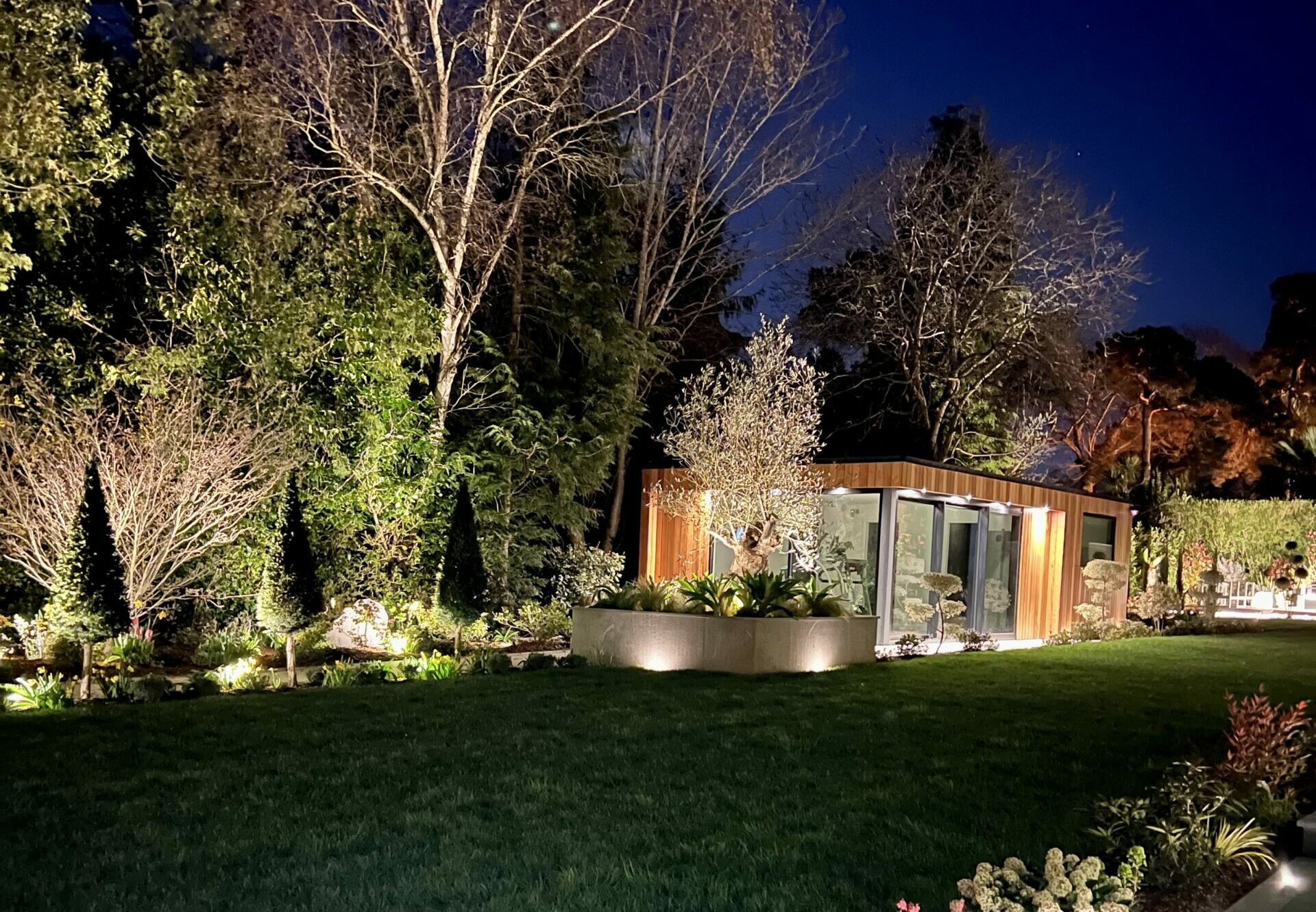Exterior night view of a Vivid Green garden gym with a flat roof, grey framed corner glass sliding doors, surrounded by a tiled patio, trees in the background, and lush foliage